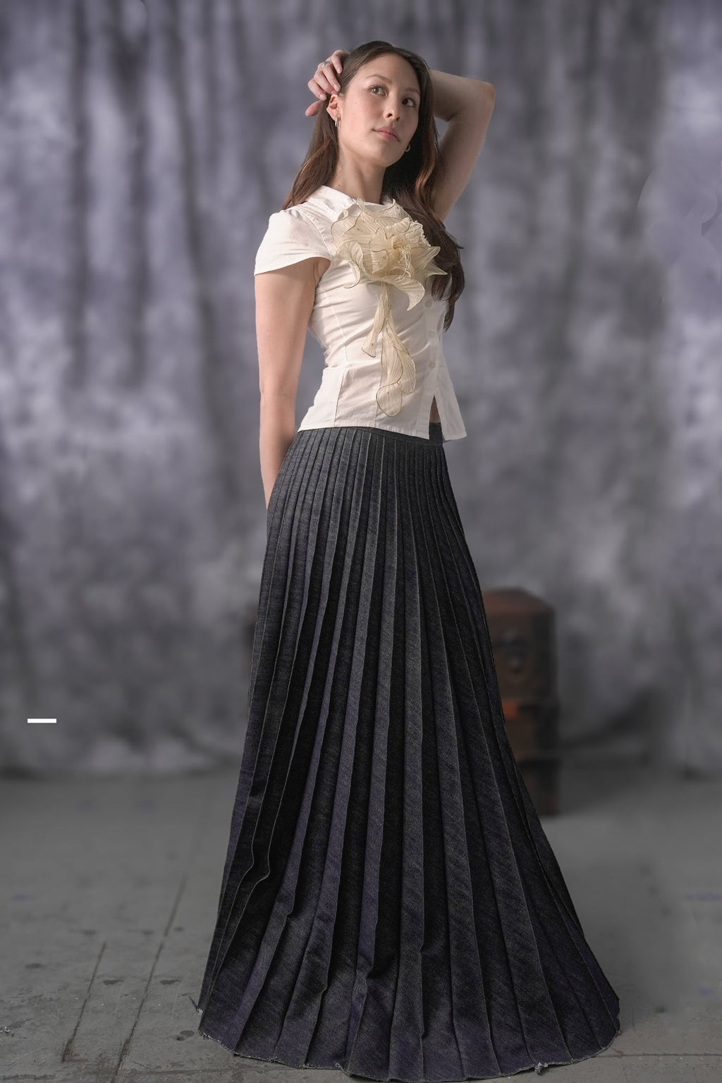 Woman wearing a white top with a large flower and a black pleated skirt against a gray background