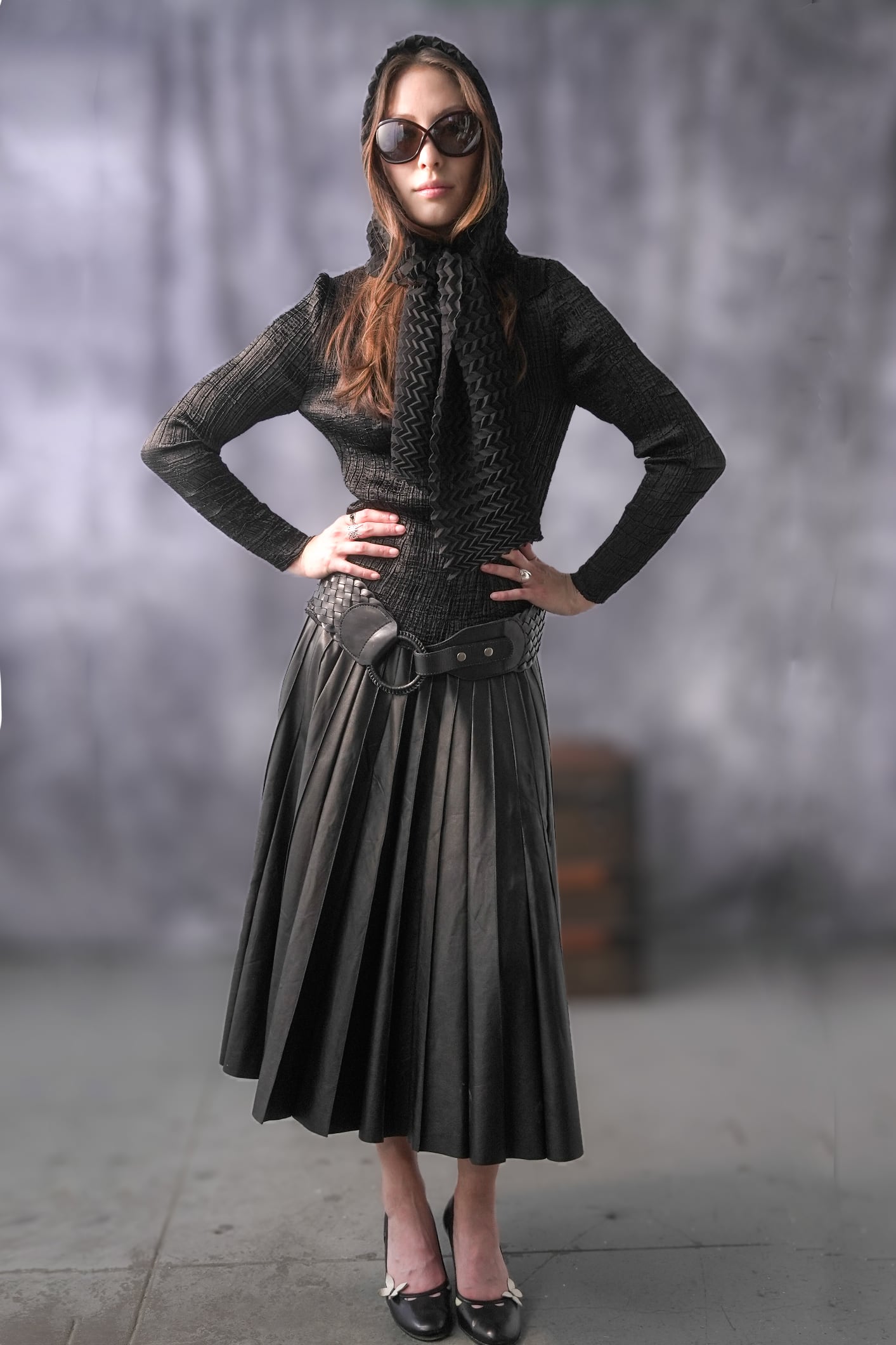 Woman wearing a black outfit with a hood and sunglasses against a gray background